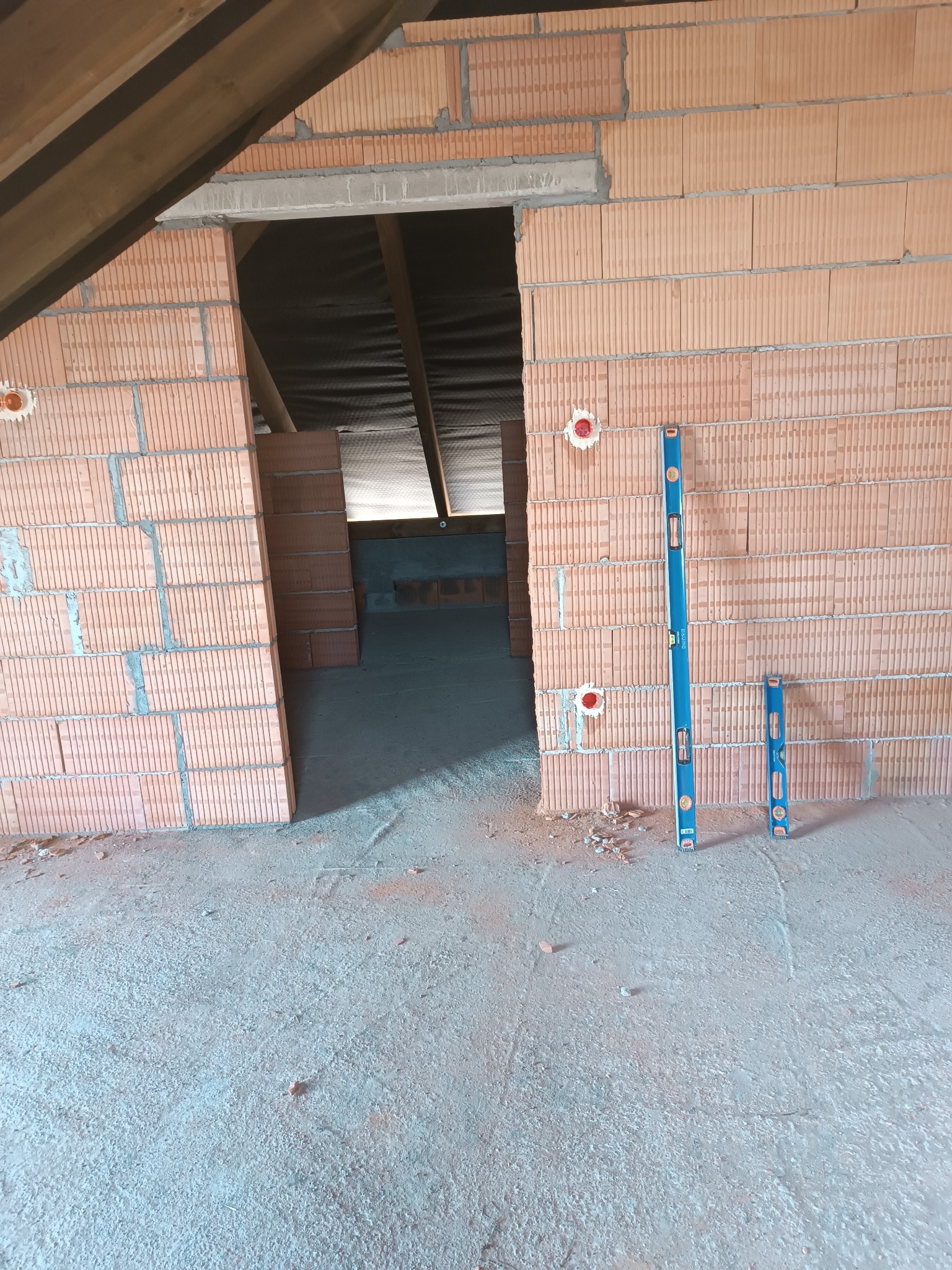 Surowy stan poddasza: ściany z cegieł dziurawki z otworami na puszki elektryczne, poziomice przy ścianie, widok na dalszą część poddasza przez otwór w ścianie.