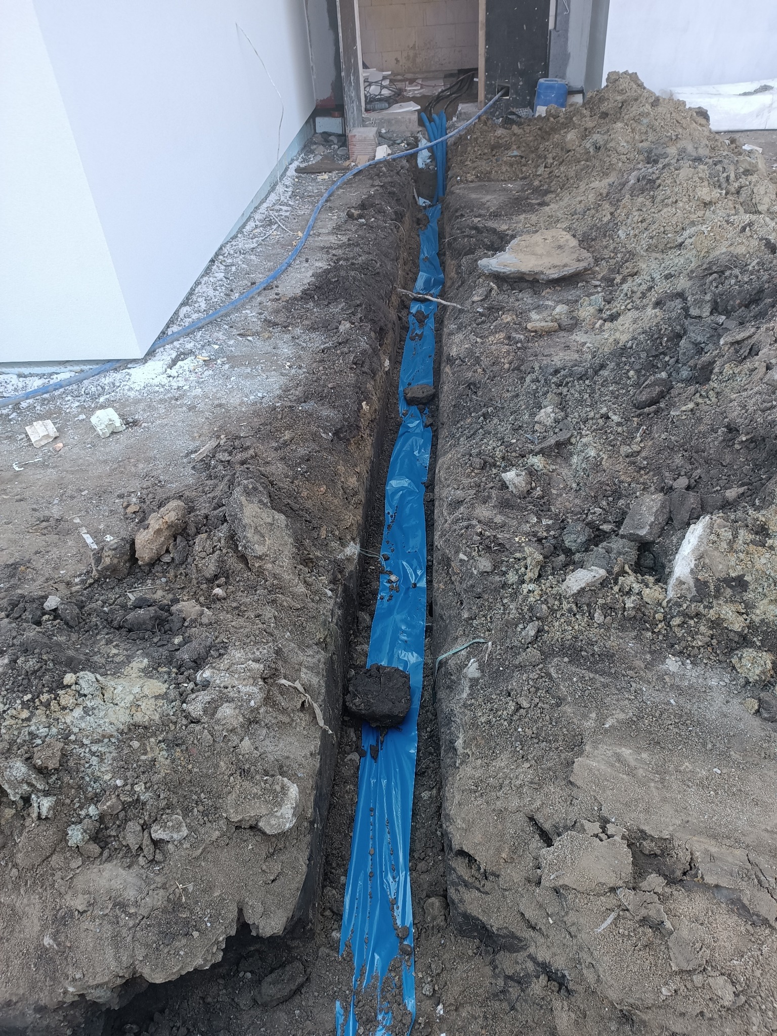 Wykop w ziemi z niebieską folią ochronną i rurami, prawdopodobnie instalacja elektryczna w trakcie układania przed zasypaniem. Widoczne przewody i ziemia.