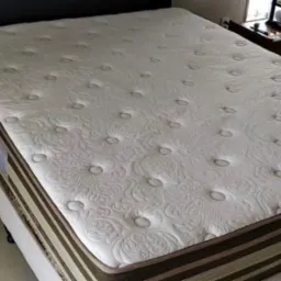 Materac z kremową tapicerką w kwiatowe wzory i guzikami, widoczny fragment ramy łóżka i stolika nocnego w tle.