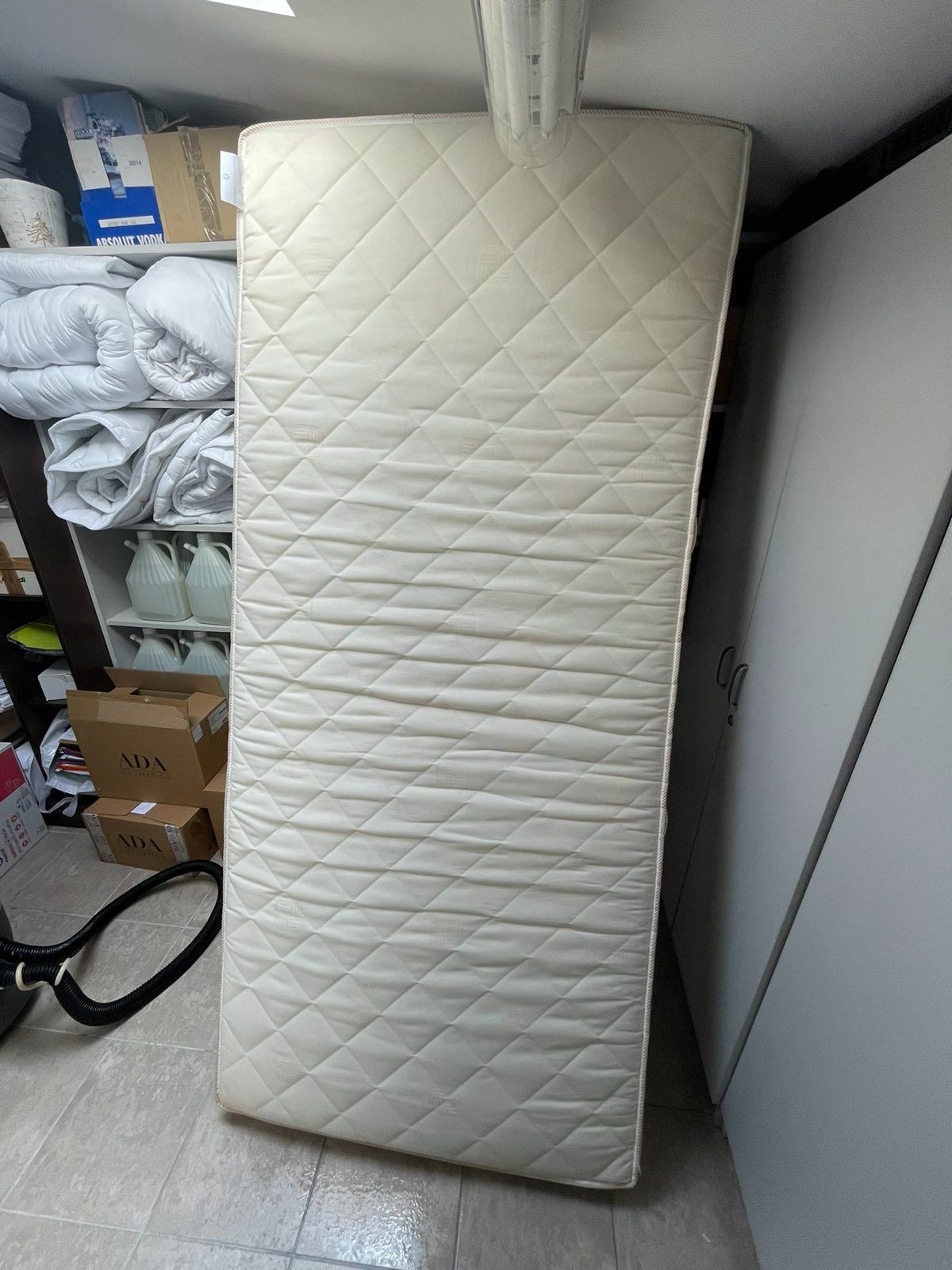 Pionowo ustawiony materac z pikowanym wzorem w romby, oparty o białą szafę, z widocznymi elementami otoczenia: rury odkurzacza, półki z pościelą i pudełkami.