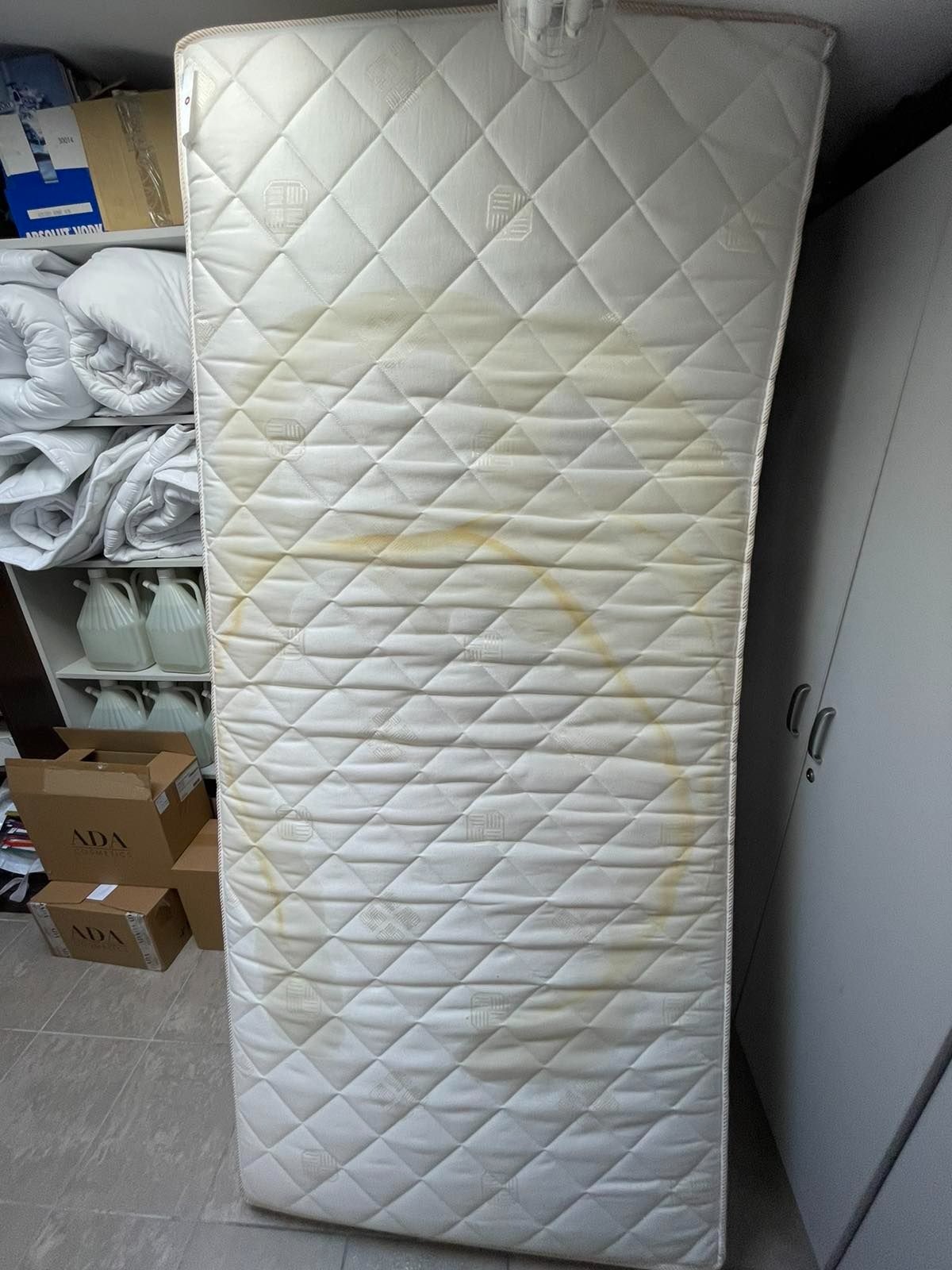 Biały materac z widocznymi, okrągłymi, żółtymi plamami na tle regału z pościelą i kartonami.