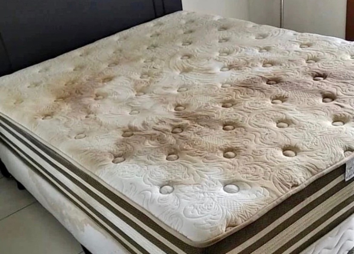 Mocno zabrudzony materac z widocznymi plamami na powierzchni, umieszczony na łóżku z ciemnym zagłówkiem.