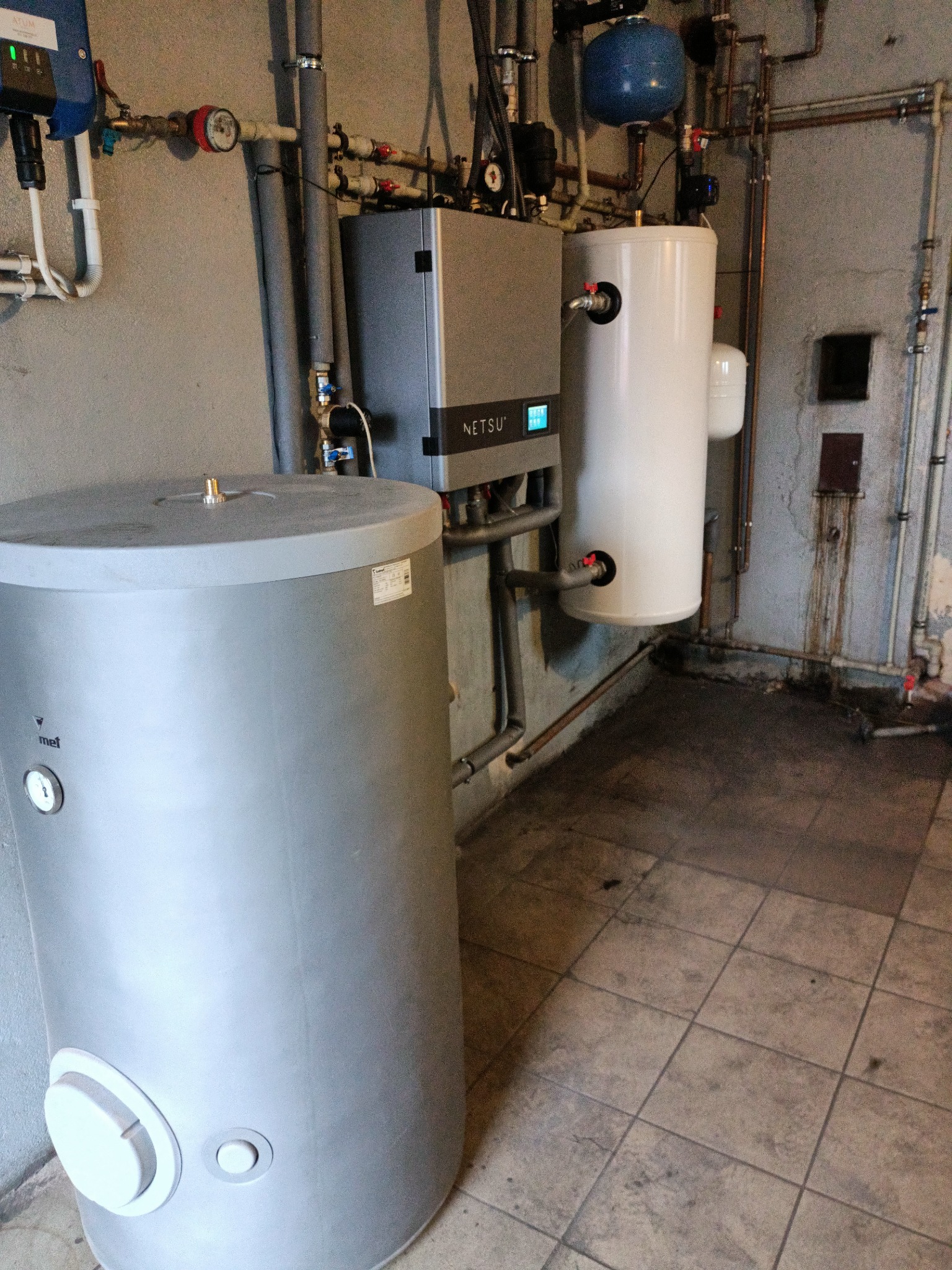 Szary zasobnik ciepłej wody użytkowej MET i biały bufor ciepła obok kotła NETSU w piwnicy, widoczne rury instalacji grzewczej na ścianie z płytkami.