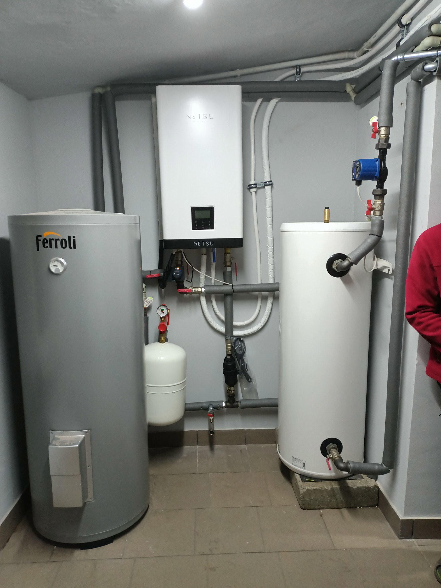 Instalacja grzewcza w kotłowni: szary zasobnik Ferroli, biały zasobnik i wiszący biały moduł Netsu z wyświetlaczem, rury w izolacji, zawory, manometry, mały biały zbiornik wyrównawczy, płytki...