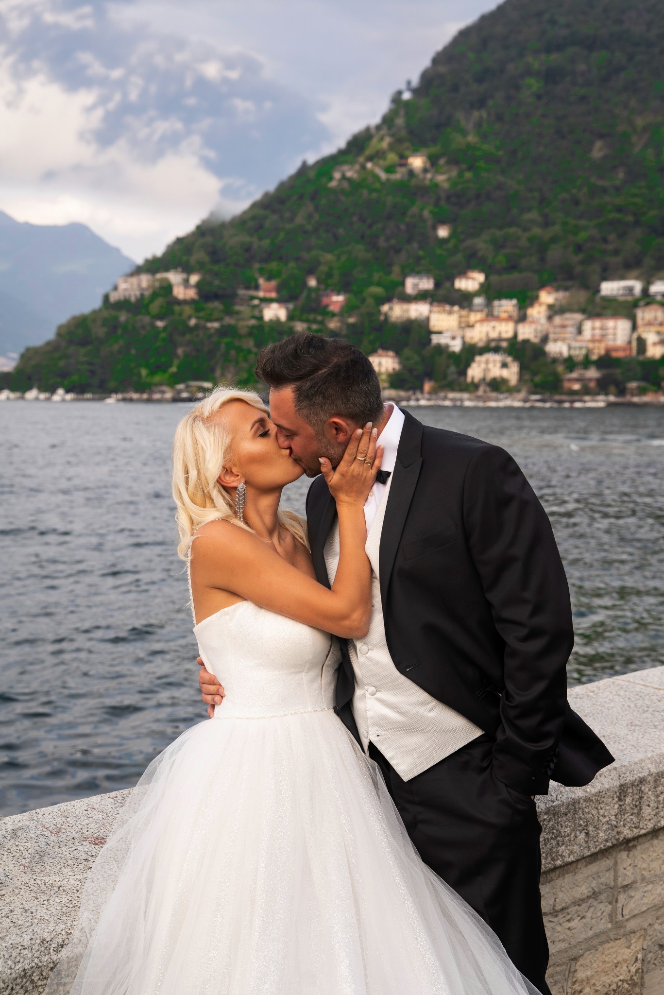Pocałunek nowożeńców na tle jeziora Como i zielonych wzgórz z zabudowaniami, panna młoda w sukni bez ramiączek, pan młody w czarnym smokingu z białą kamizelką.