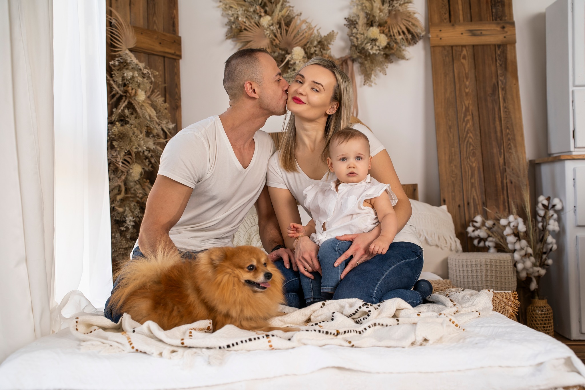 Rodzinna sesja zdjęciowa w studio: tata całuje mamę w policzek, trzymając na kolanach córeczkę, obok leży pomeranian, w tle drewniane dekoracje i suszone kwiaty.