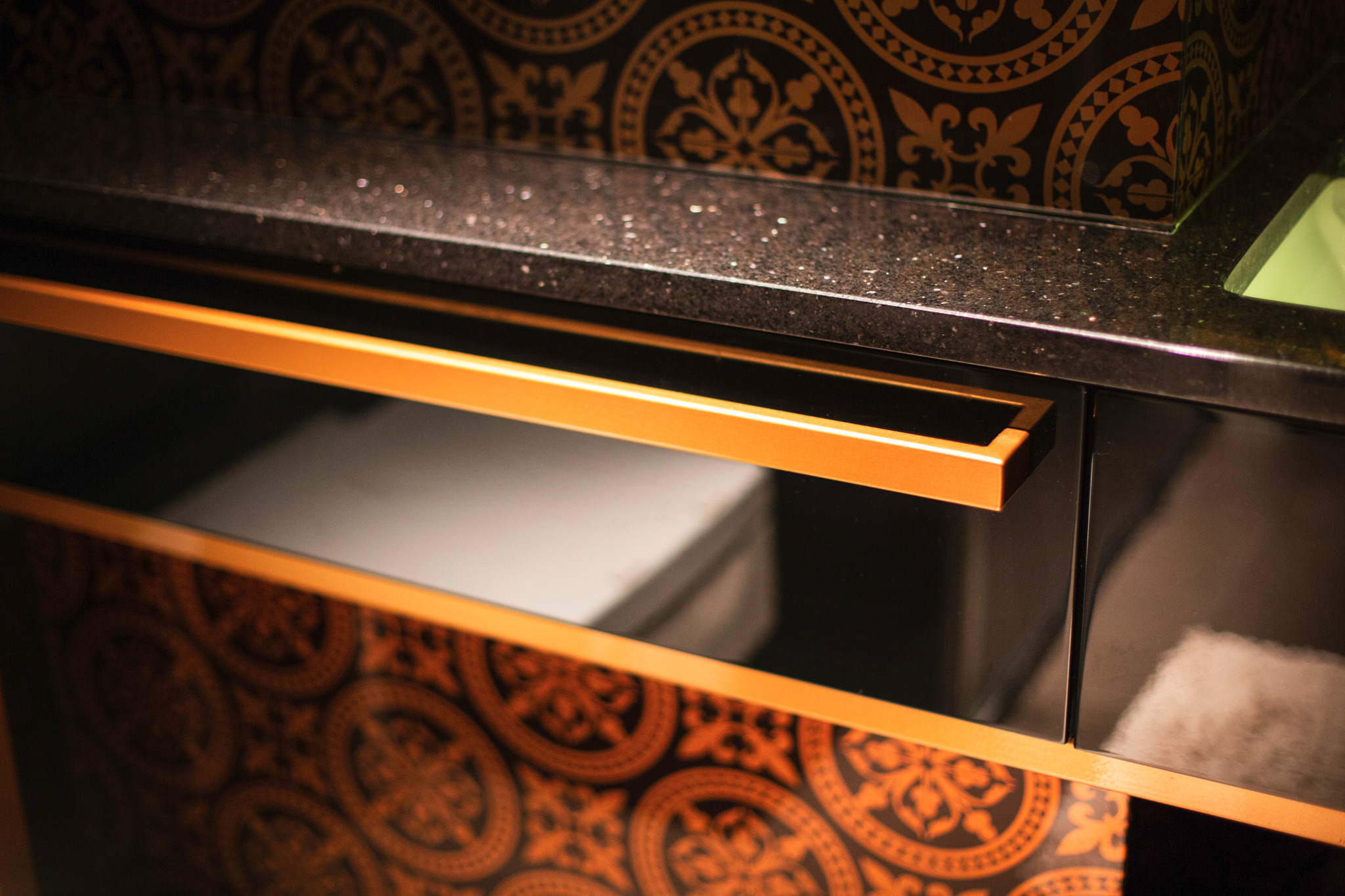 Szklana balustrada z czarnym blatem i złotymi zdobieniami, widoczny fragment umywalki