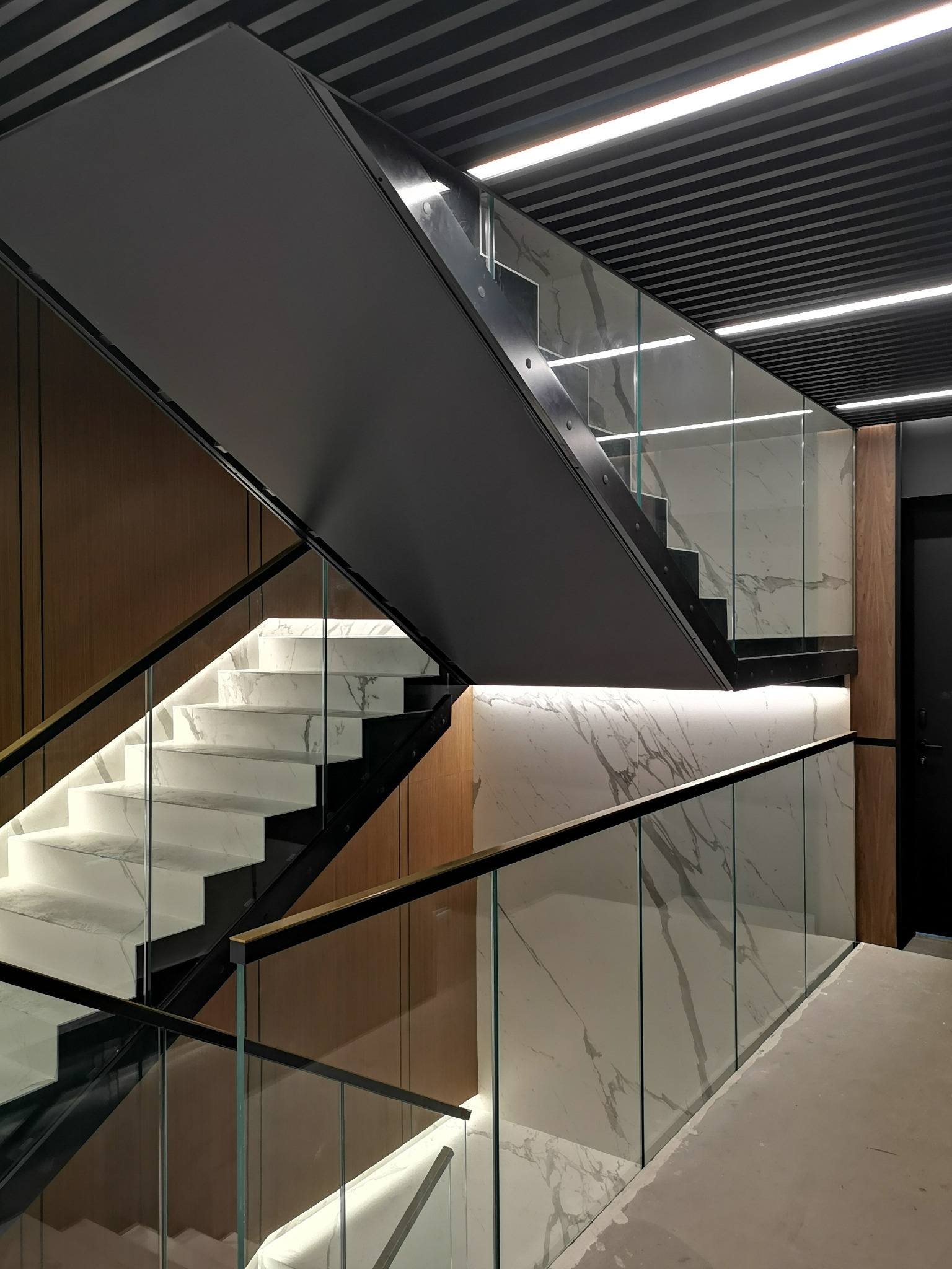 Schody wewnętrzne z marmurowymi stopniami i szklaną balustradą, podświetlone od spodu, z czarną konstrukcją i drewnianymi panelami na ścianach, widok z dołu.