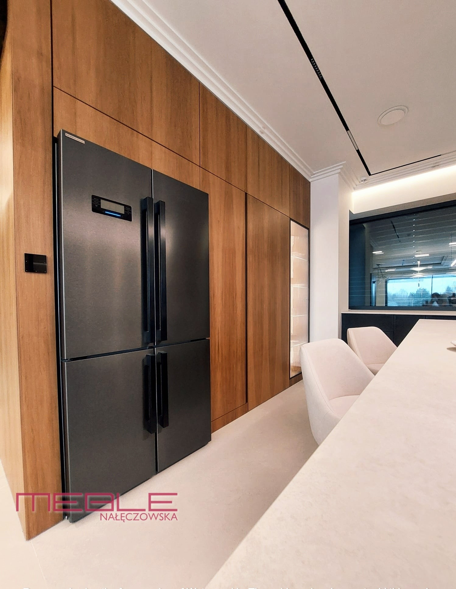 Nowoczesna kuchnia z zabudową z drewnianych paneli, zintegrowana z dużą, czarną, czterodrzwiową lodówką. W tle widoczny stół i krzesła w jasnych barwach.