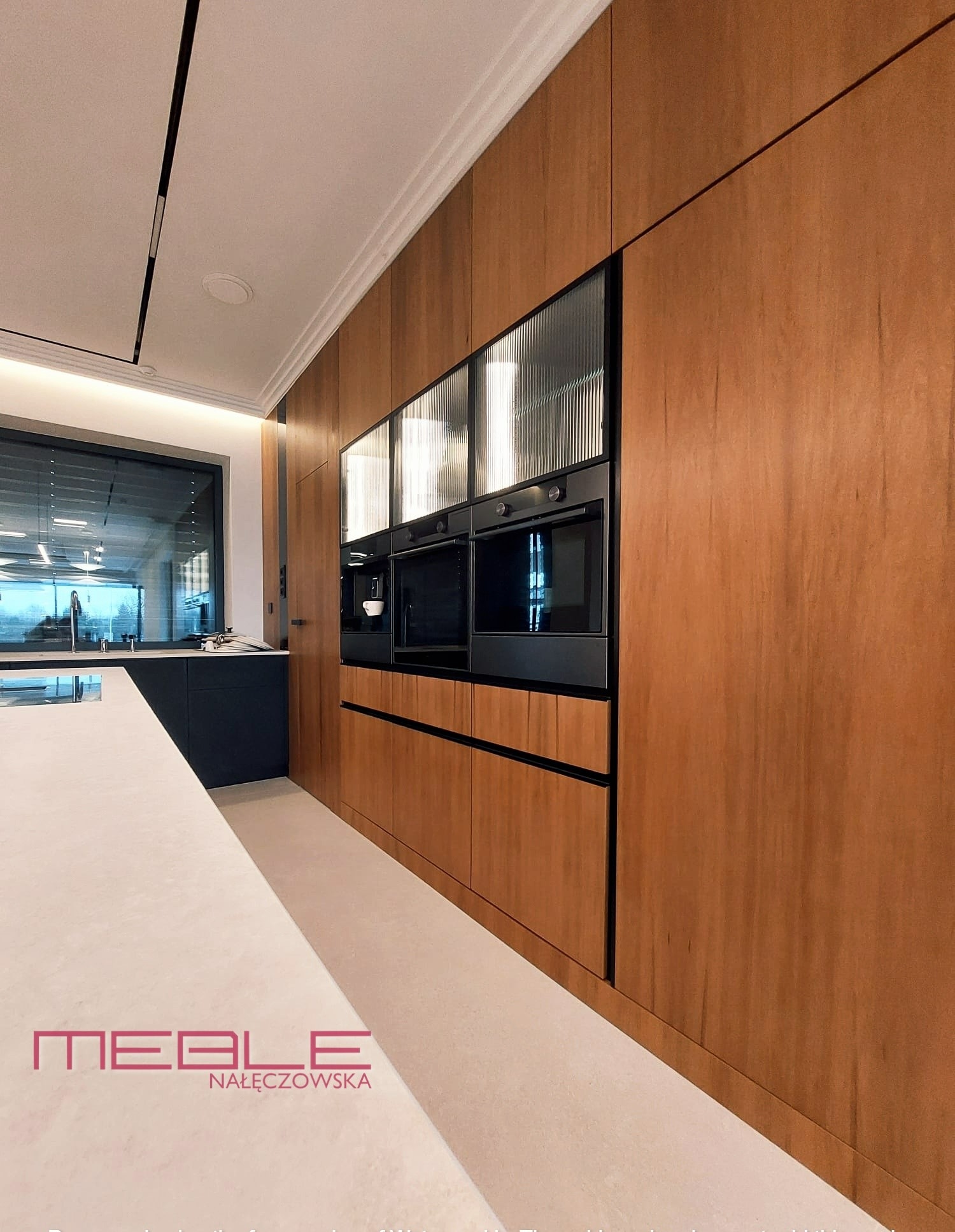 Nowoczesna kuchnia z drewnianymi frontami, czarnymi akcentami i zabudowanymi urządzeniami, widok z boku na logo firmy na blacie.