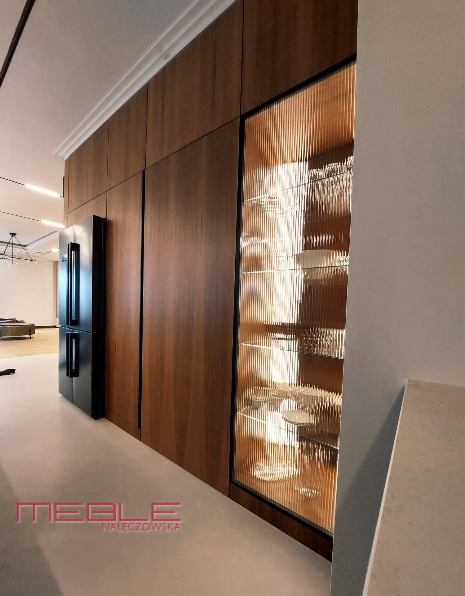 Ciemne, drewniane szafy z czarnymi detalami, w tym przeszklona witryna z ryflowanym szkłem, eksponująca zastawę stołową, zintegrowane z czarną lodówką w nowoczesnym wnętrzu.