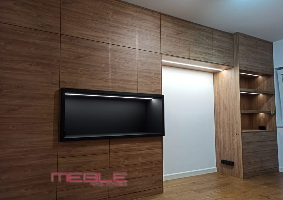Drewniana zabudowa ściany z szafkami i wnęką na telewizor, oświetlenie LED w górnej części wnęki i półek.