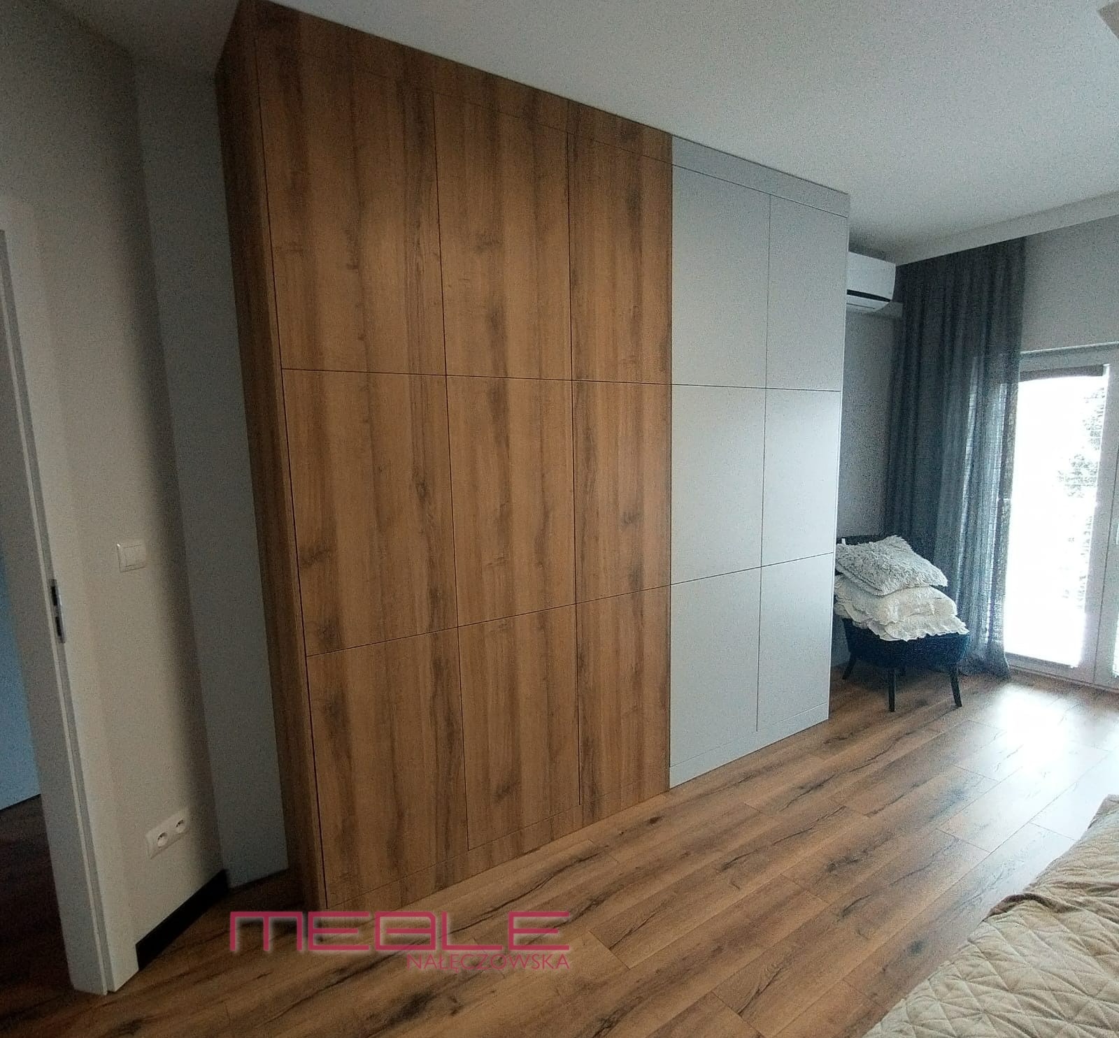 Duża szafa z frontami w kolorze drewna i szarego matu, widoczna w sypialni z drewnianą podłogą i krzesłem z poduszkami.