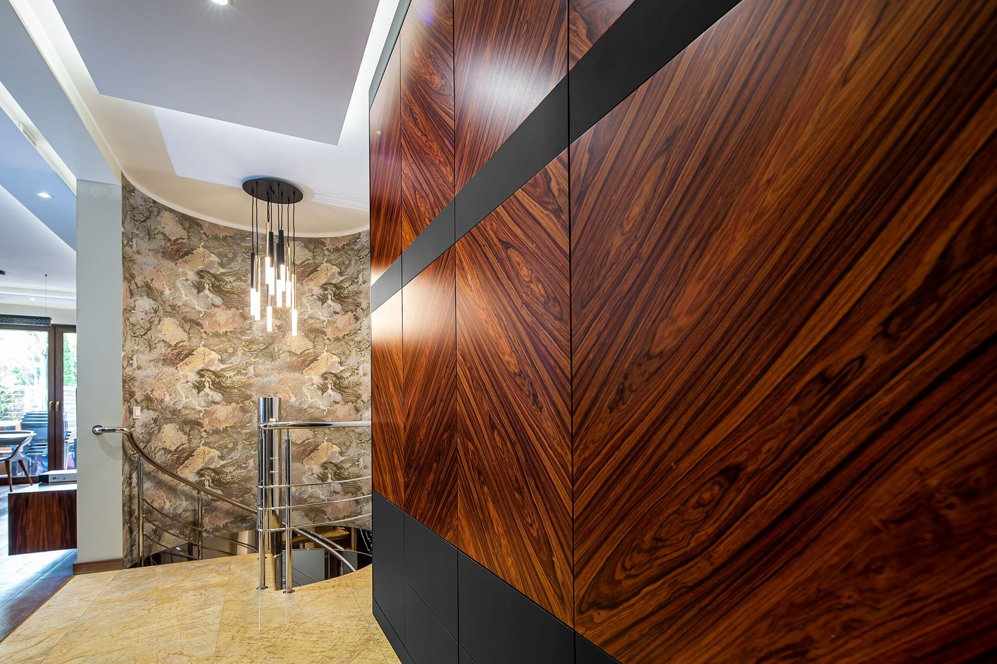 Wnętrze domu z dużą szafą wnękową z egzotycznego drewna o wzorze chevron, czarne akcenty, marmurowa podłoga i schody z metalową balustradą.
