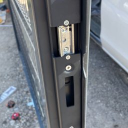 BS BudSerwis - Szczegółowe ujęcie zamka w panelowym ogrodzeniu, widoczne śruby mocujące i mechanizm blokujący, samochód w tle.