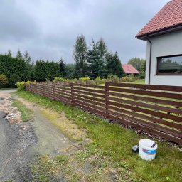 BS BudSerwis - Długie, poziome, drewniane ogrodzenie w kolorze brązowym, widoczne od strony drogi gruntowej i przylegającego trawnika, z wiadrem farby i pędzlem na trawie, dom w tle.