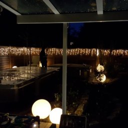 BS BudSerwis - Nocny widok na oświetlony ogród z drewnianym ogrodzeniem, dekorowany girlandami świetlnymi i kulistymi lampami ogrodowymi. Częściowo zadaszony taras z meblami.