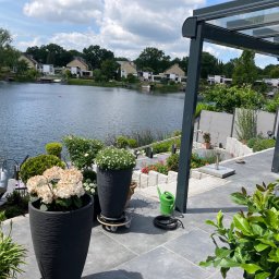 BS BudSerwis - Nowoczesny taras z szarych płyt, z widokiem na jezioro. Donice z kwiatami, zieleń, zadaszenie tarasu. W tle zabudowa jednorodzinna, błękitne niebo z chmurami.