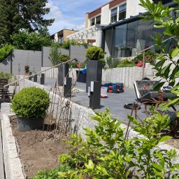 BS BudSerwis - Nowoczesny taras z szarej kostki, obrzeża z kamienia, minimalistyczne ogrodzenie panelowe i roślinność. Widoczne narzędzia sugerują etap prac wykończeniowych.