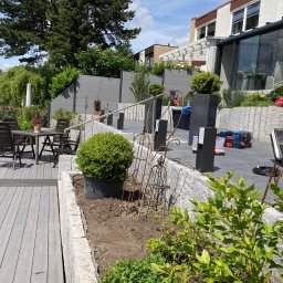 BS BudSerwis - Nowoczesny taras z szarej deski kompozytowej, otoczony kamienną rabatą i minimalistycznym ogrodzeniem panelowym. Widoczne narzędzia i skrzynki narzędziowe.