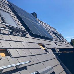 BS BudSerwis - Montaż paneli słonecznych i okien dachowych na dachu z szarej dachówki, widoczne elementy mocujące i fragment niekompletnego pokrycia. Błękitne niebo w tle.