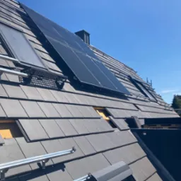 Montaż paneli słonecznych i okien dachowych na dachu z szarej dachówki, widoczne elementy mocujące i fragment niekompletnego pokrycia. Błękitne niebo w tle.