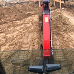 BS BudSerwis - Czerwona koparka w wykopie z piasku, widok z kabiny operatora na teren budowy z drewnianymi znacznikami, w tle widoczne rury i ogrodzenie.