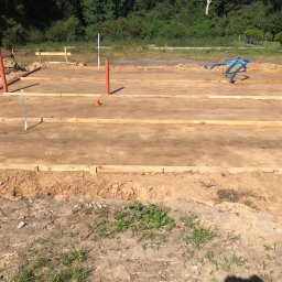 BS BudSerwis - Przygotowanie terenu pod budowę tarasu drewnianego: widoczne drewniane obramowanie, rury instalacyjne. W tle zieleń drzew i krzewów.