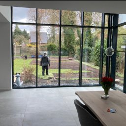 BS BudSerwis - Widok z wnętrza domu na układanie trawy z rolki w ogrodzie. Przez duże okno widać pracownika, rozłożone rolki trawy i częściowo zagospodarowany teren.