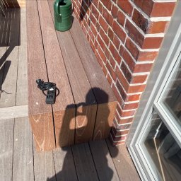 BS BudSerwis - Nowoczesny taras drewniany z kostką, zielona konewka przy ceglanej ścianie, cień osoby. Listwa zasilająca leży na tarasie. Słoneczny dzień.