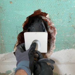 BS BudSerwis - Instalacja puszki elektrycznej w ścianie z odsłoniętą cegłą, widoczne ręce w roboczych rękawicach. Ściana pomalowana na turkusowo z widocznymi śladami wcześniejszych warstw.