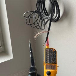 BS BudSerwis - Elektryk w niebieskich rękawicach trzyma miernik napięcia przy przewodach wystających ze ściany. Widoczny fragment okna w tle.