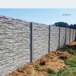BS BudSerwis - Panelowe ogrodzenie betonowe o fakturze kamienia, z widocznymi słupkami. Ujęcie z poziomu gruntu, w tle zieleń drzew i błękit nieba. Widoczne logo producenta.