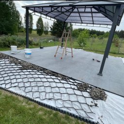 BS BudSerwis - Budowa tarasu z szarej kostki pod zadaszeniem. Widoczna warstwa drenażowa z geokraty, kamieni i obrzeża. W tle zieleń ogrodu i drabina. Etap prac przed zasypaniem.