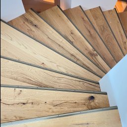 BS BudSerwis - Spiralne, drewniane schody z metalowymi wykończeniami, widziane z góry, z fragmentem osoby w skarpetkach na dolnym stopniu. Jasne oświetlenie, minimalistyczny design.