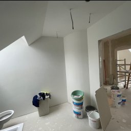 BS BudSerwis - Poddasze w trakcie malowania na biało. Widoczne wiadra z farbą, drabina w przejściu do innego pomieszczenia, instalacja elektryczna w suficie i łazienka w tle.