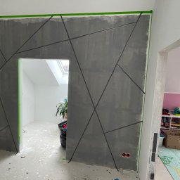 BS BudSerwis - Ściana po malowaniu z geometrycznym wzorem w szarościach, zabezpieczona taśmą malarską. Widoczne dwa pomieszczenia w tle.