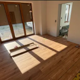 Montaż paneli podłogowych w Olsztynie. Częściowo ułożona podłoga z widoczną izolacją i narzędziami, intensywne światło słoneczne wpadające przez okno.