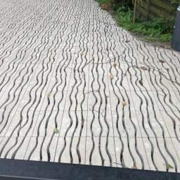 BS BudSerwis - Szara nawierzchnia z falistymi szczelinami, przypominająca ułożoną kostkę brukową, z elementami roślinnymi i koszem na śmieci w tle. Nietypowy wzór kostki.