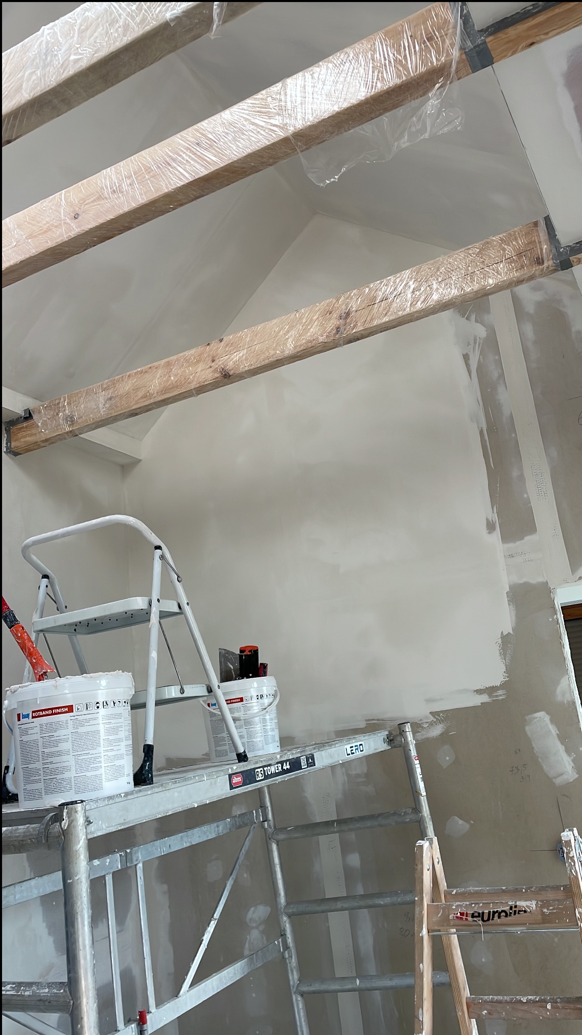 Skośna ściana po szpachlowaniu, widoczna z rusztowania z wiadrami gładzi. Belki stropowe owinięte folią ochronną.