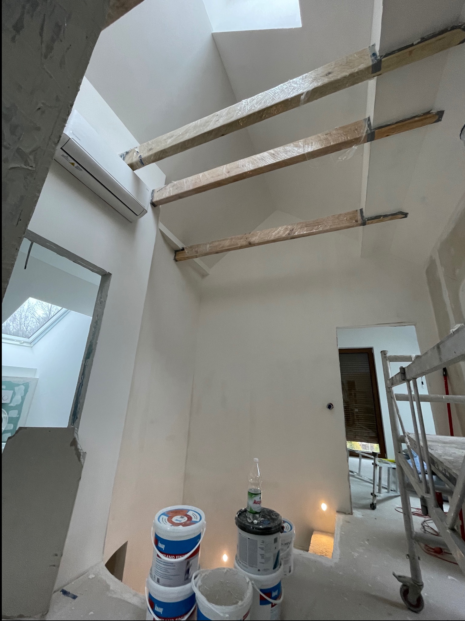 Wnętrze w trakcie wykańczania: sufit z belkami, ściany przygotowane do malowania, widoczne puszki z farbą i rusztowanie. Przez okno widać drzewa.