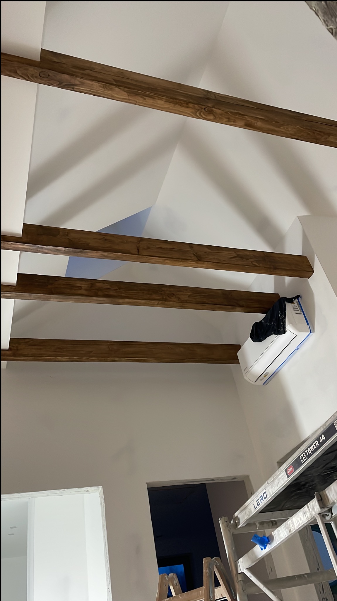 Wnętrze z drewnianymi belkami stropowymi i klimatyzacją zabezpieczoną folią, widok z dołu, fragment rusztowania. Prace wykończeniowe w trakcie realizacji.