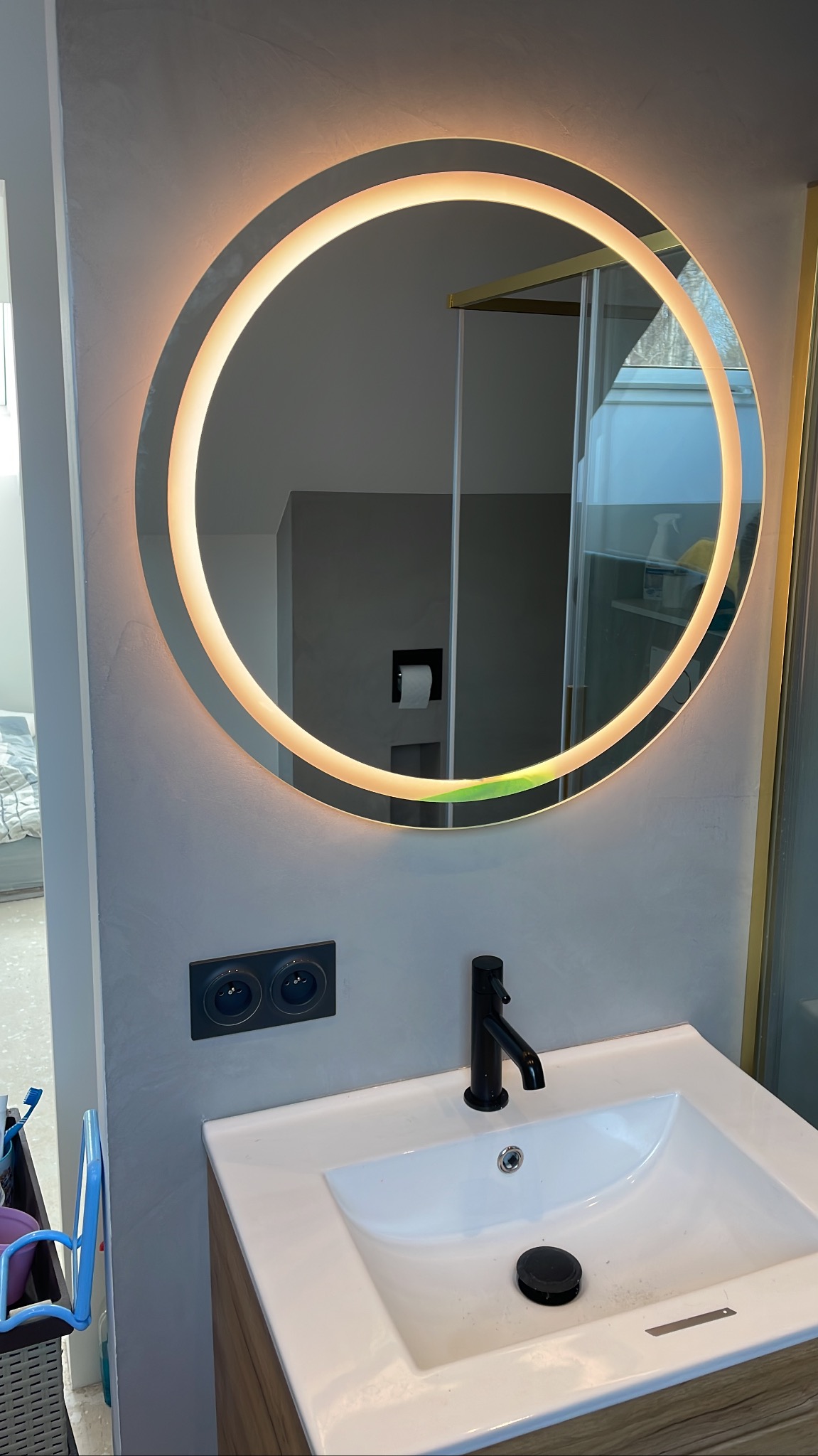 Nowoczesna łazienka: biała umywalka z czarną baterią, okrągłe lustro z podświetleniem LED i szare ściany z widocznymi gniazdkami elektrycznymi.