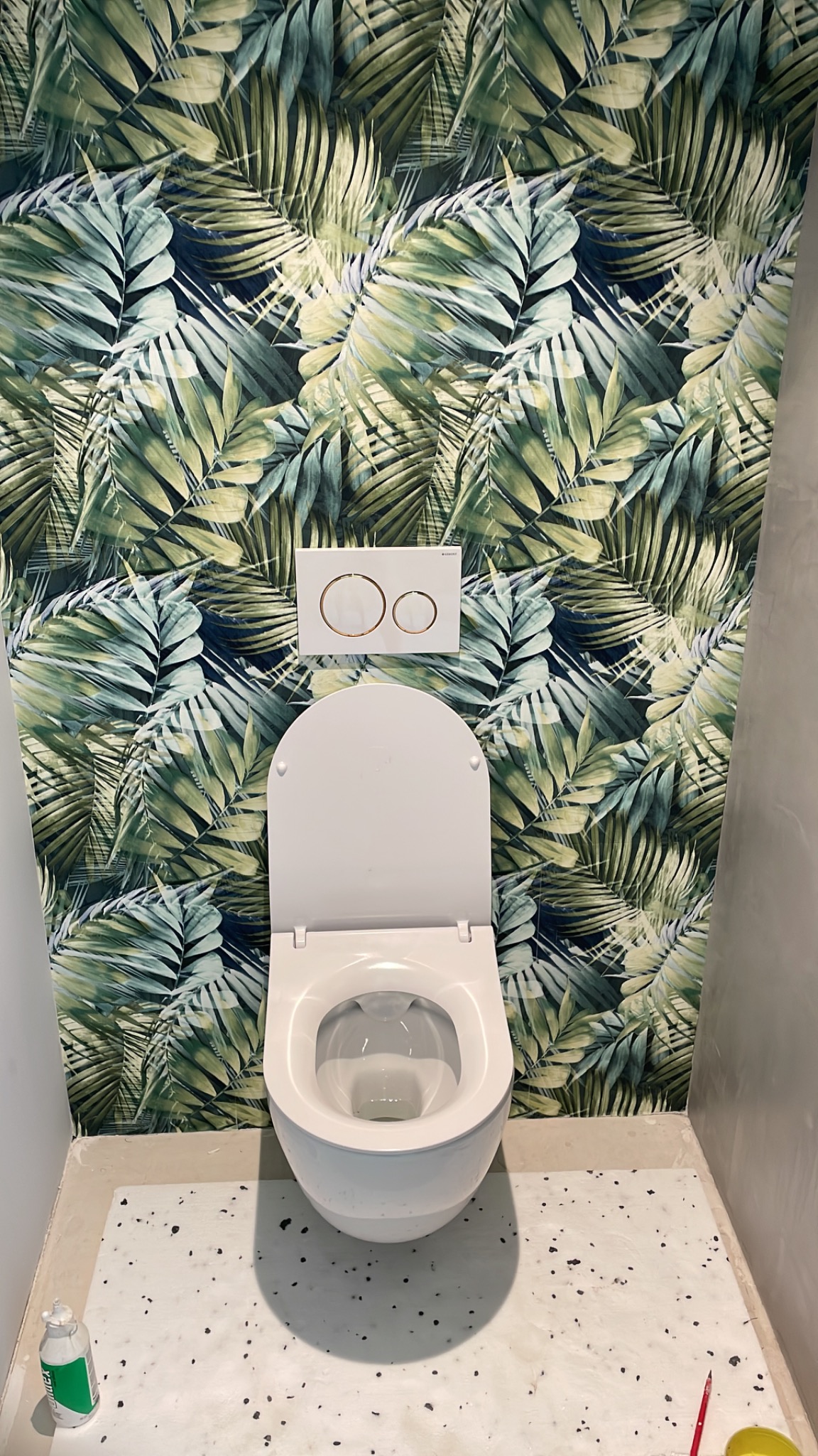 Biała toaleta z deską na tle ściany w tropikalne liście. Widoczne przyciski spłukiwania. Na podłodze białe płytki z czarnymi plamkami i resztki po remoncie.