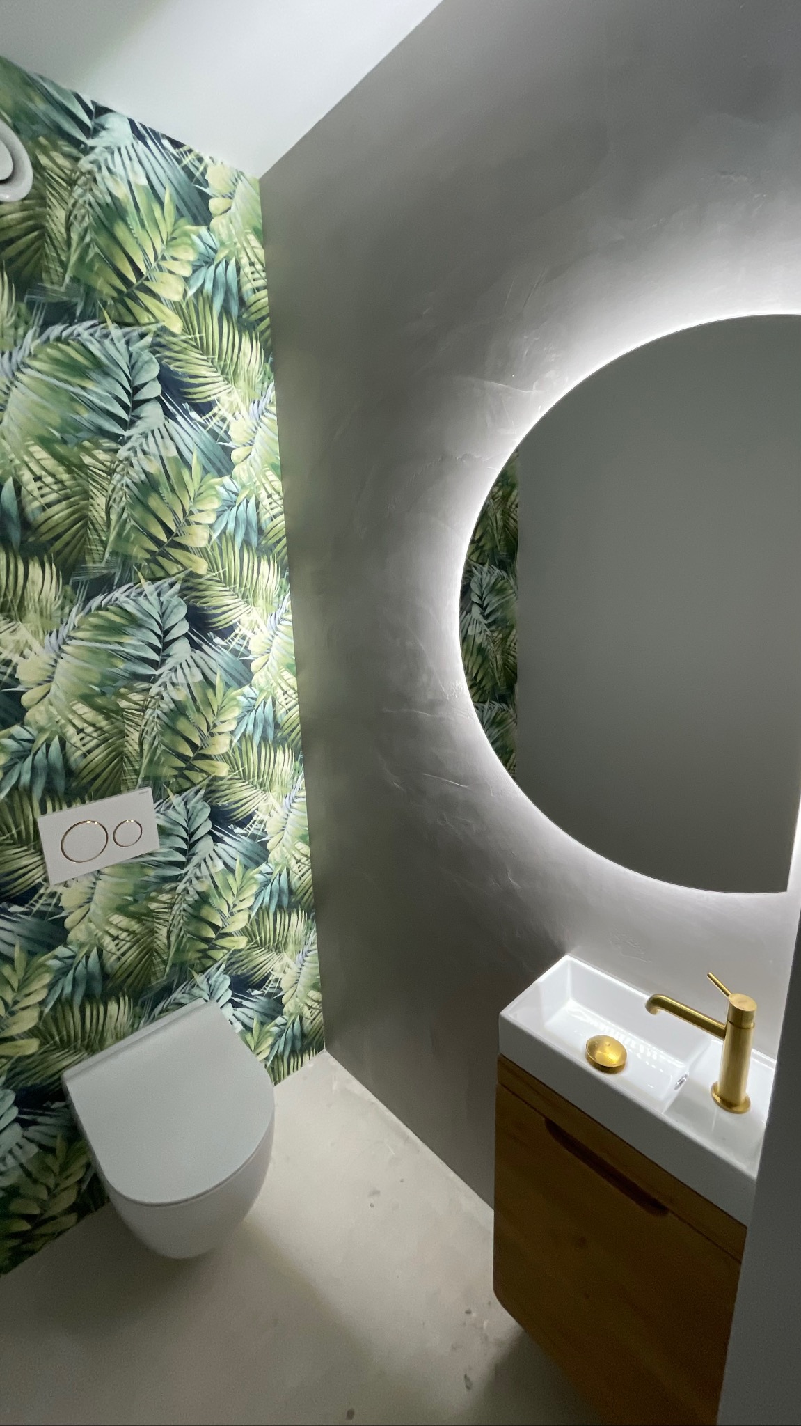 Nowoczesna łazienka z toaletą, umywalką z szafką i okrągłym lustrem z podświetleniem LED. Ściana z motywem tropikalnych liści dodaje egzotycznego charakteru.