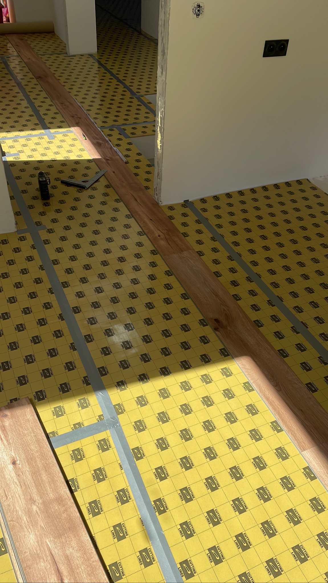 Układanie paneli podłogowych na żółtym podkładzie z narzędziami. Widoczne fragmenty podłogi i ściany w jasnym wnętrzu. W trakcie montażu.