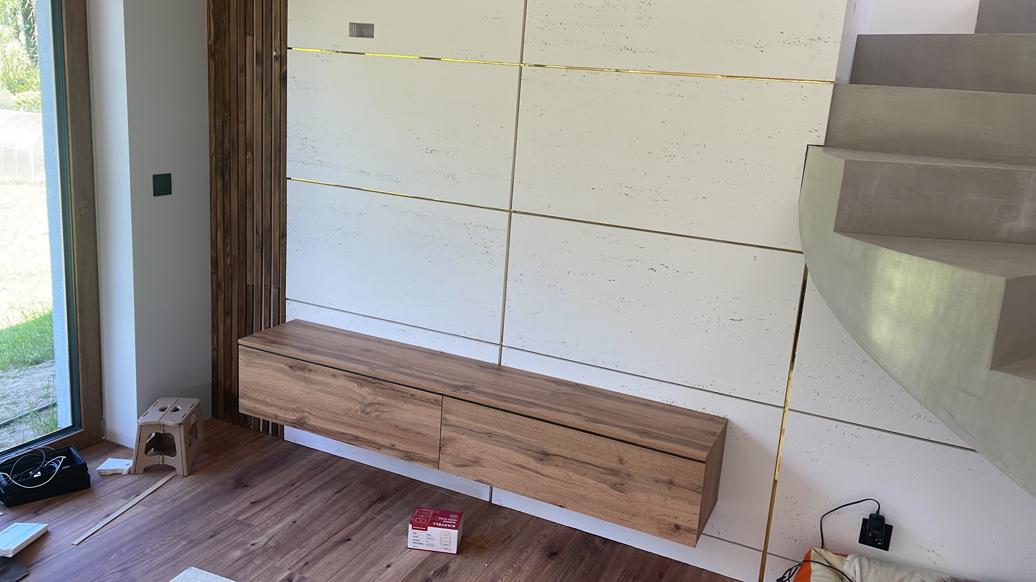 Nowoczesne wnętrze z drewnianą szafką RTV pod ścianą z białych paneli z złotymi akcentami, drewnianą ścianką działową i betonowymi schodami.