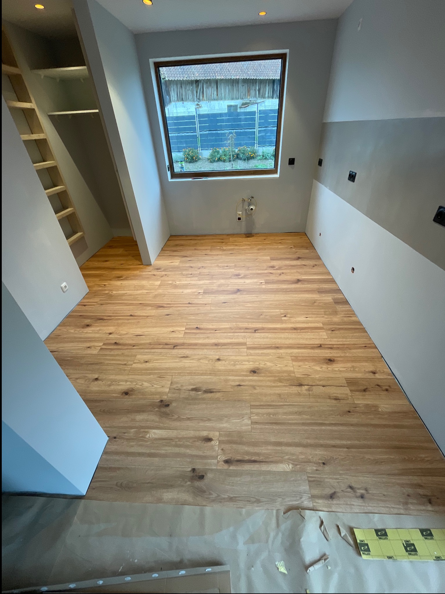 Jasne panele podłogowe ułożone w pokoju na poddaszu z oknem i wnęką, widok z góry. Część podłogi zabezpieczona folią. Widoczne gniazdka elektryczne.