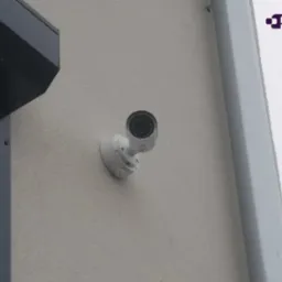 Biała kamera kopułkowa CCTV zamontowana na jasnej, teksturowanej ścianie zewnętrznej budynku, widoczny fragment dachu i rynny.