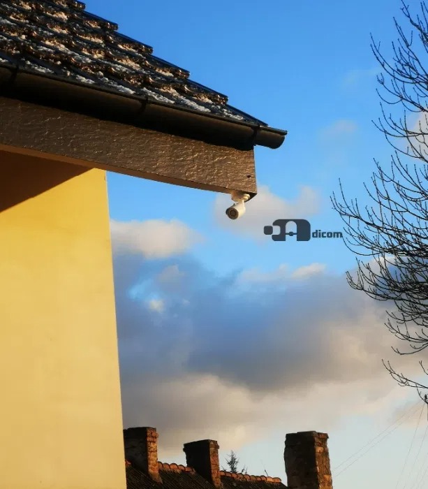 Kamera kopułkowa zamontowana pod okapem budynku jednorodzinnego, widok z dołu na tle błękitnego nieba z chmurami.